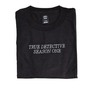 Puede incluir: Camiseta negra con el texto blanco "TRUE DETECTIVE SEASON ONE". La camiseta está doblada y parece estar hecha de un material suave. Tiene cuello redondo y mangas cortas.