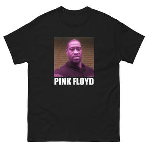 Puede incluir: Camiseta negra con un retrato de un hombre y el texto blanco "PINK FLOYD". El retrato está enmarcado en un rectángulo sobre un fondo de pared de ladrillo. La camiseta es de cuello redondo.