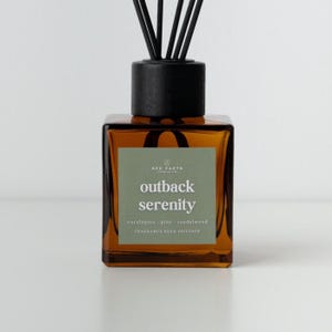 May include: An amber glass fragrance reed diffuser with a black wooden cap and reeds. The label reads "Outback Serenity" with eucalyptus, pine, and sandalwood fragrance notes. The product is a fragrance reed diffuser.