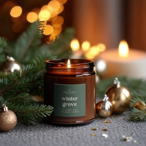 May include: A brown glass jar candle with a lit wick and a green label that reads Red Earth Candle Co. winter grove fir needle, pine, spearmint, amber. The candle is surrounded by green pine branches and small gold ornaments.