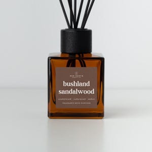 May include: An amber glass fragrance reed diffuser with black reeds. The label reads "bushland sandalwood" with scent notes. The diffuser is from Red Earth Candle Co. and sits on a white surface.