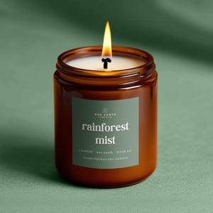 May include: A burning soy candle in a brown glass jar. The label says "rainforest mist" with "rainfall, wet earth, fresh air" below. The candle is hand-poured.