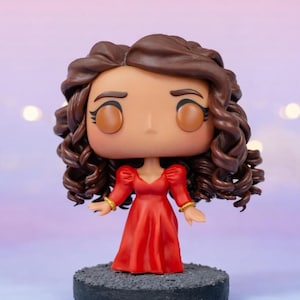 May include: A stylized vinyl collectible figure featuring a character with voluminous dark brown curly hair, wearing a long-sleeved red dress with gold-colored cuffs. The figure has large solid brown eyes, defined eyebrows, and stands on a circular base.
