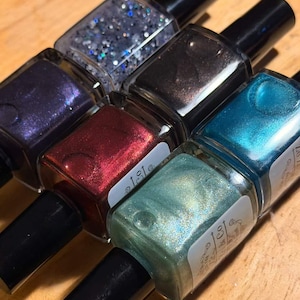 May include: A collection of six nail polish bottles in various colors, including purple, red, teal, and silver glitter. Each bottle has a black cap and a clear glass body, showcasing the polish color.
