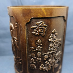 Op de afbeelding: Bronskleurige cilindrische container met gebeeldhouwde bloemen- en scriptontwerpen. De container heeft een licht uitlopende bovenkant en een gedetailleerd reliëf van bloemen en Chinese karakters. De algehele esthetiek is antiek.