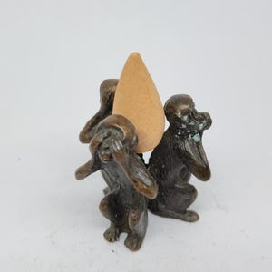 Può includere: Scultura in bronzo di tre scimmie nella posa "Non vedere il male, non sentire il male, non parlare il male", che tengono un cono di incenso marrone. Le scimmie hanno dettagli e una finitura invecchiata. Un oggetto decorativo.