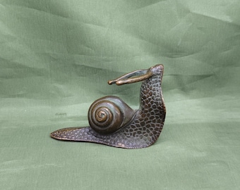 May include: A bronze snail sculpture with a detailed shell and textured body. The snail's head is raised, and its antennae are extended. The sculpture is set against a green background, highlighting the bronze color and intricate design.
