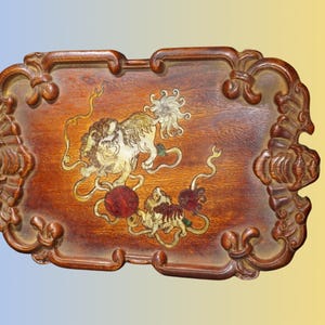 May include: A rectangular wooden serving tray with a dark brown finish and ornate, carved edges. The center of the tray features an inlaid design depicting two mythical lion-like creatures playing with a red ball, rendered in light and dark wood tones.