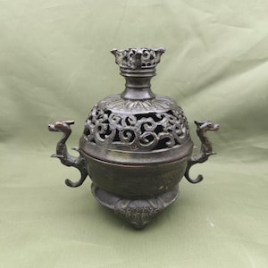 May include: A bronze-colored incense burner with intricate openwork and dragon-shaped handles. The burner has a domed lid and a decorative base, showcasing an antique aesthetic. The overall design suggests a traditional or Asian-inspired theme.