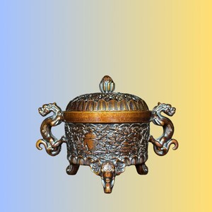 May include: A bronze-colored metal incense burner featuring a removable lid with a finial, two side handles shaped like dragon heads, and three legs designed as elephant heads. The body is decorated with intricate relief carvings of floral motifs.