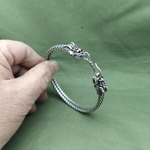 May include: A silver bracelet with a dragon head design at each end. The bracelet is made of woven metal and has a hook clasp. The dragon heads are detailed with scales and facial features. The bracelet is shown against a green background.