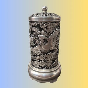 May include: A silver-colored, cylindrical container with a lid and a round base. The container features intricate carvings of a bird in flight amidst clouds. The lid is topped with a small, round knob. The design is detailed and ornate.