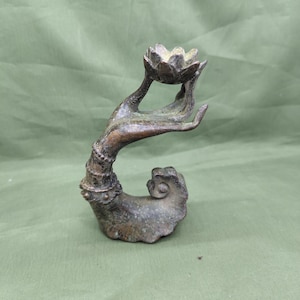 May include: Bronze sculpture of a hand holding a lotus flower. The hand is intricately detailed with a curved base and ornate wrist adornments. The lotus flower is in full bloom, and the sculpture has a weathered, antique appearance.