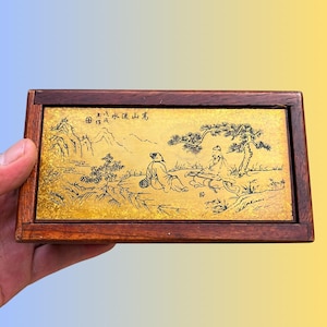 Könnte beinhalten: Eine rechteckige Holzschatulle mit einer goldfarbenen Metalleinlage auf dem Deckel. Die Einlage zeigt eine schwarze Tuschezeichnung von zwei Figuren, die unter einer Kiefer bei Bergen sitzen, mit chinesischen Schriftzeichen oben.
