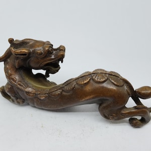 May include: Bronze-colored figurine of a mythical creature, possibly a dragon or a similar beast. The sculpture is detailed, with visible scales and a fierce expression. The creature is in a resting pose.