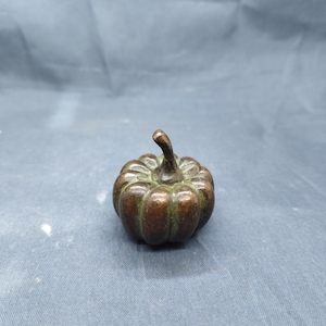May include: A small, bronze-colored pumpkin figurine with a detailed ribbed texture and a curved stem. The miniature pumpkin is a decorative item, likely for fall or Halloween.