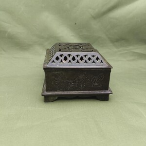 May include: A dark bronze rectangular incense burner with a decorative floral design on the sides. The top features a lattice pattern and cut-out designs. The burner sits on a small, rectangular base.
