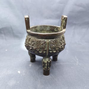 Może przedstawiać: Brązowy, trójnożny palnik do kadzidła z dwoma uchwytami. Naczynie posiada skomplikowane, wytłaczane wzory wokół korpusu. Palnik ma zaokrąglony kształt i ciemną, postarzaną patynę, co sugeruje, że jest to antyk lub przedmiot w stylu vintage. Projekt jest prawdopodobnie pochodzenia azjatyckiego.