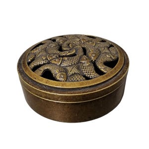 Puede incluir: Un recipiente redondo de color bronce con una tapa que presenta un relieve de peces. El recipiente tiene una abertura circular en la tapa, que revela el diseño de los peces. El recipiente probablemente se utiliza para almacenamiento o fines decorativos.