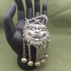 May include: A silver-colored pendant necklace featuring a detailed, stylized lion's head with a wide grin and dangling chain accents. The pendant is held by a black hand against a green background.