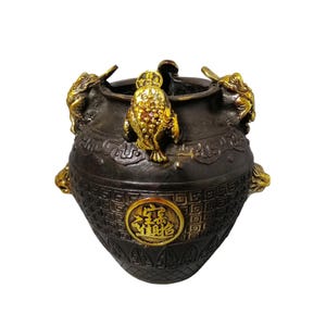 Puede incluir: Una olla decorativa de color bronce con detalles dorados. La olla presenta detalles ornamentados, incluyendo patrones en relieve y criaturas míticas doradas. Un emblema dorado con caracteres chinos está en el frente.