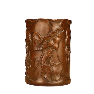 May include: A cylindrical, brown wooden brush pot with carved relief details. The design features a fish, lotus flowers, and leaves. The pot has a slightly irregular top edge and a smooth, polished finish.