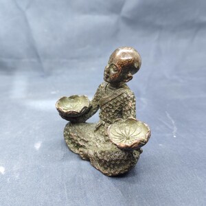 May include: A bronze-colored figurine of a seated monk holding two lotus-shaped bowls. The monk is depicted with a shaved head and detailed robes. The statue has an aged, antique appearance.