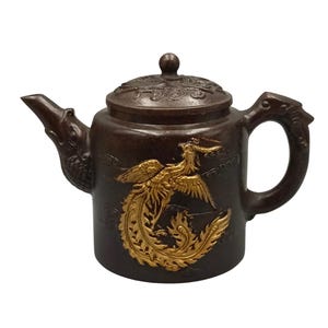 May include: A brown teapot with a gold phoenix design. The teapot has a curved spout and handle, and a lid with a small knob. The phoenix is depicted with its wings spread, and the teapot has intricate details.