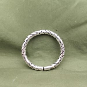 May include: A silver-colored bangle bracelet with a twisted rope design. The bracelet is circular and has a clasp. The background is a solid olive green color.