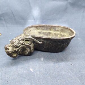 May include: A bronze-colored, oval-shaped bowl with a dragon head handle. The bowl has a weathered appearance, with intricate details on the dragon's face and body. The bowl's surface shows signs of age and use, suggesting it is an antique.