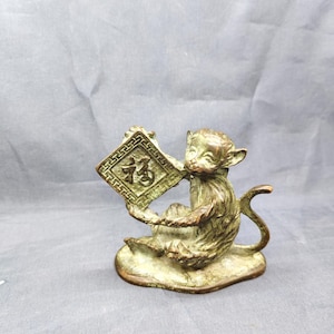 May include: Bronze monkey figurine holding a square plaque with Chinese characters. The monkey is seated on a base, with a curled tail. The plaque has a decorative border and the characters are in the center. The figurine has a weathered appearance.