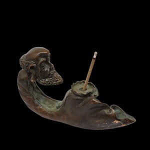 May include: A bronze incense burner depicting a seated figure with a detailed face and beard. The figure holds a small bowl with an incense stick. The sculpture has a dark brown patina with hints of green.