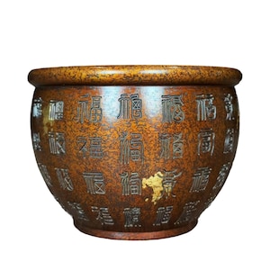 May include: A round, brown ceramic pot with embossed Chinese characters. The pot has a textured surface and a gold-colored emblem. The rim is a darker shade of brown. The pot is likely used for decorative purposes.