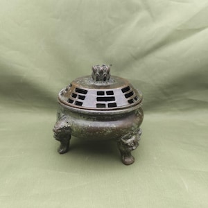 May include: Bronze incense burner with a domed lid featuring geometric cutouts and a floral finial. The burner rests on three legs with detailed lion-like faces. The overall design is antique and ornate.