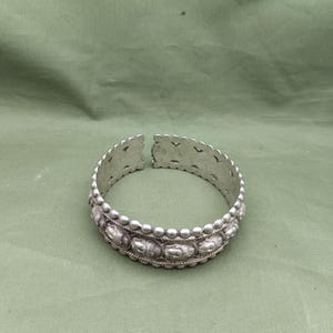Puede incluir: Brazalete de puño plateado con un diseño abierto. El brazalete presenta un patrón detallado de pequeñas cuentas y formas ovaladas. Los bordes tienen un diseño decorativo festoneado. El brazalete se muestra sobre una superficie verde.