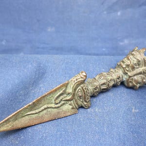 May include: An antique bronze dagger with detailed carvings. The handle features faces and ornate designs, while the blade is a simple, sharp triangle. The dagger has a weathered appearance, suggesting it is a vintage item.