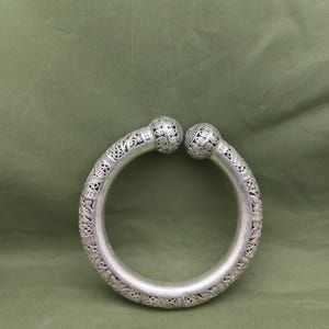 Puede incluir: Brazalete de puño plateado con patrones intrincados y detallados. El brazalete tiene forma redondeada con extremos decorativos en forma de bola. El diseño presenta motivos florales y geométricos, creando una estética vintage o antigua.