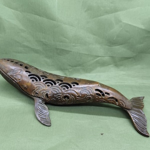 以下が含まれることがあります： 背中に波と渦の複雑な切り抜き模様が施されたブロンズ製のクジラの置物。クジラは詳細な尾とひれを持っています。この置物は装飾品で、緑色の表面に置かれています。