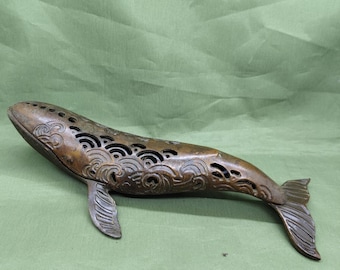 May include: Bronze whale figurine with intricate wave and swirl cut-out designs along its back. The whale has a detailed tail and fins, and is resting on a green surface. The figurine is a decorative item.