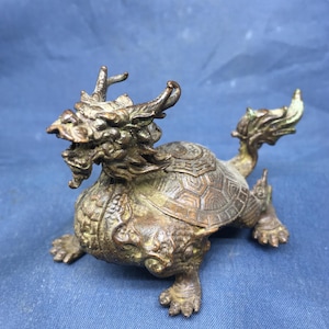 May include: A bronze-colored figurine depicting a mythical creature with a dragon's head, horns, and a turtle-like shell. The detailed sculpture features intricate patterns and textures, with four clawed feet and a curled tail. The figurine is a decorative item.
