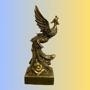May include: A metallic gold-colored brass sculpture of a phoenix in flight, featuring intricate feather detailing on the wings and long tail. The mythical bird is perched atop a stylized cloud base, which rests on a solid, rectangular block pedestal.