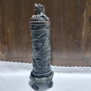 May include: A dark gray, cylindrical incense burner with intricate dragon carvings. The burner features a dragon figure on the lid and a detailed base. The design is ornate and the overall aesthetic is antique.
