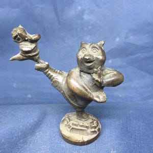 Puede incluir: Una figura de bronce de un personaje parecido a un oso en una pose juguetona, sosteniendo una figura más pequeña. La base es redonda con un diseño detallado. El estilo general sugiere un tema caprichoso o de dibujos animados.