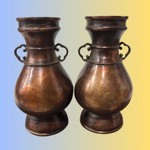 May include: Two antique copper vases with a bulbous body, flared neck, and decorative handles. The vases have a dark, aged patina, suggesting they are vintage or antique pieces. The design is symmetrical, with a wide base and a narrow neck.