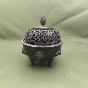 Puede incluir: Un quemador de incienso de color bronce con un diseño calado decorativo en la tapa y el cuerpo. El quemador tiene tres patas pequeñas y un diseño floral en la base. La tapa está rematada con un pequeño adorno en forma de capullo.