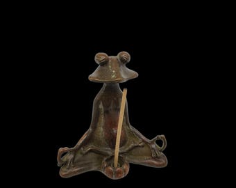 May include: Bronze frog figurine in a meditative pose, with arms crossed and legs folded. The frog holds a wooden incense stick in a small bowl. The figurine has a dark brown color and is set against a black background.
