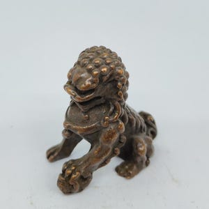 May include: A small, bronze-colored statue of a mythical lion-like creature. The figurine has a detailed mane, a fierce expression, and is posed in a seated position. The sculpture is likely made of metal and is on a white background.