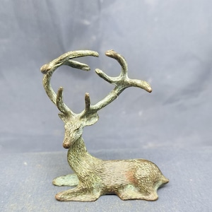 May include: A bronze deer figurine in a resting pose. The deer has large antlers and is detailed with textured fur. The figurine is a decorative item, likely for display on a shelf or table.