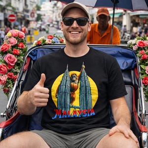 Puede incluir: Una persona con una camiseta negra con un gr&aacute;fico de las Torres Petronas y un orangut&aacute;n, con la palabra "MALAYSIA" debajo. La persona est&aacute; sentada en un rickshaw decorado, haciendo una se&ntilde;al de pulgar.