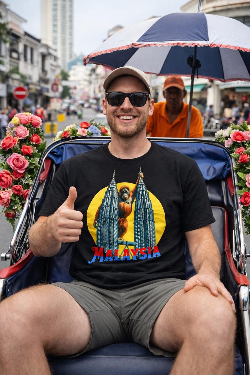 Puede incluir: Una persona con una camiseta negra con un gr&aacute;fico de las Torres Petronas y un orangut&aacute;n, con la palabra "MALAYSIA" debajo. La persona est&aacute; sentada en un rickshaw decorado, haciendo una se&ntilde;al de pulgar.